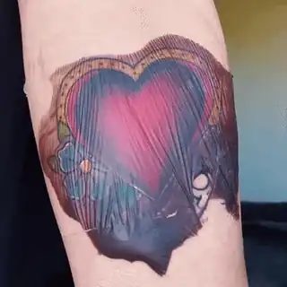Tatuaje de un corazón en el antebrazo, cubierto por una membrana transparente con líquido acumulado debajo: el film se ve arrugado y turbio, y al apretarlo con un dedo el líquido se corre.